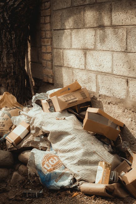 A pile of household rubbish and packaging materials, including cardboard boxes, paper bags, and plastic waste, is accumulated against a rough, weathered brick and concrete wall outdoors. The cardboard