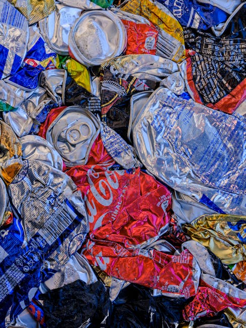 Close-up of a pile of assorted litter behind a black metal fence, including crumpled white and colored plastic bags, a torn cardboard carton with red, blue, and white markings, and various scattered p