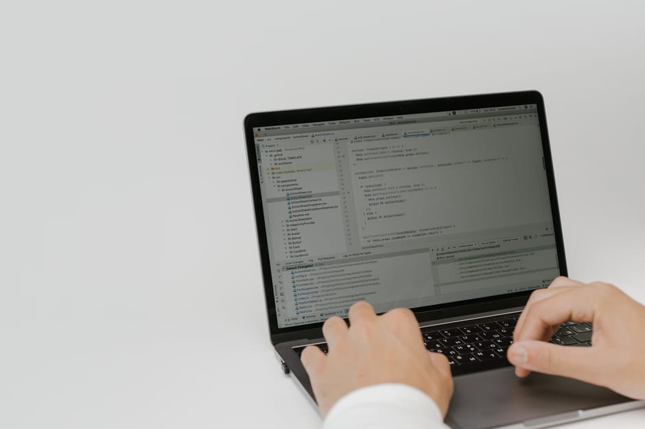 A person with short hair viewed from behind working at a desk in an office environment, focusing on a large computer monitor displaying lines of colorful programming code on a dark background. The wor