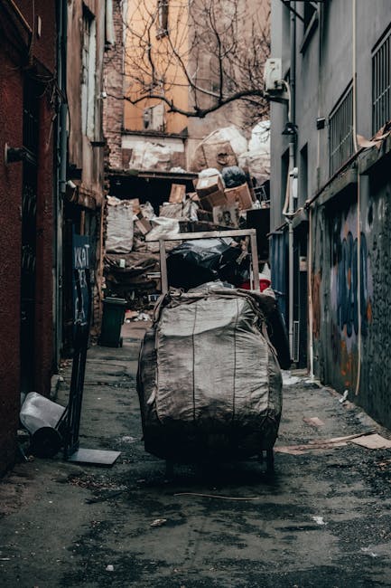 A narrow, outdoor alleyway flanked by brick and metal-clad walls, both covered in graffiti. The ground is uneven, muddy, and lined with scattered debris and dirt patches. In the foreground, there is a