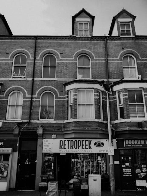 A black-and-white photograph of a multi-story brick building with a retail ground floor. The upper floors feature three evenly spaced arched windows with white frames, and two small dormer windows wit